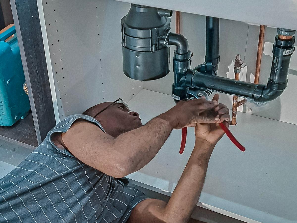 Plombier travaillant sous un évier de cuisine, réparant des tuyaux avec des outils à main.