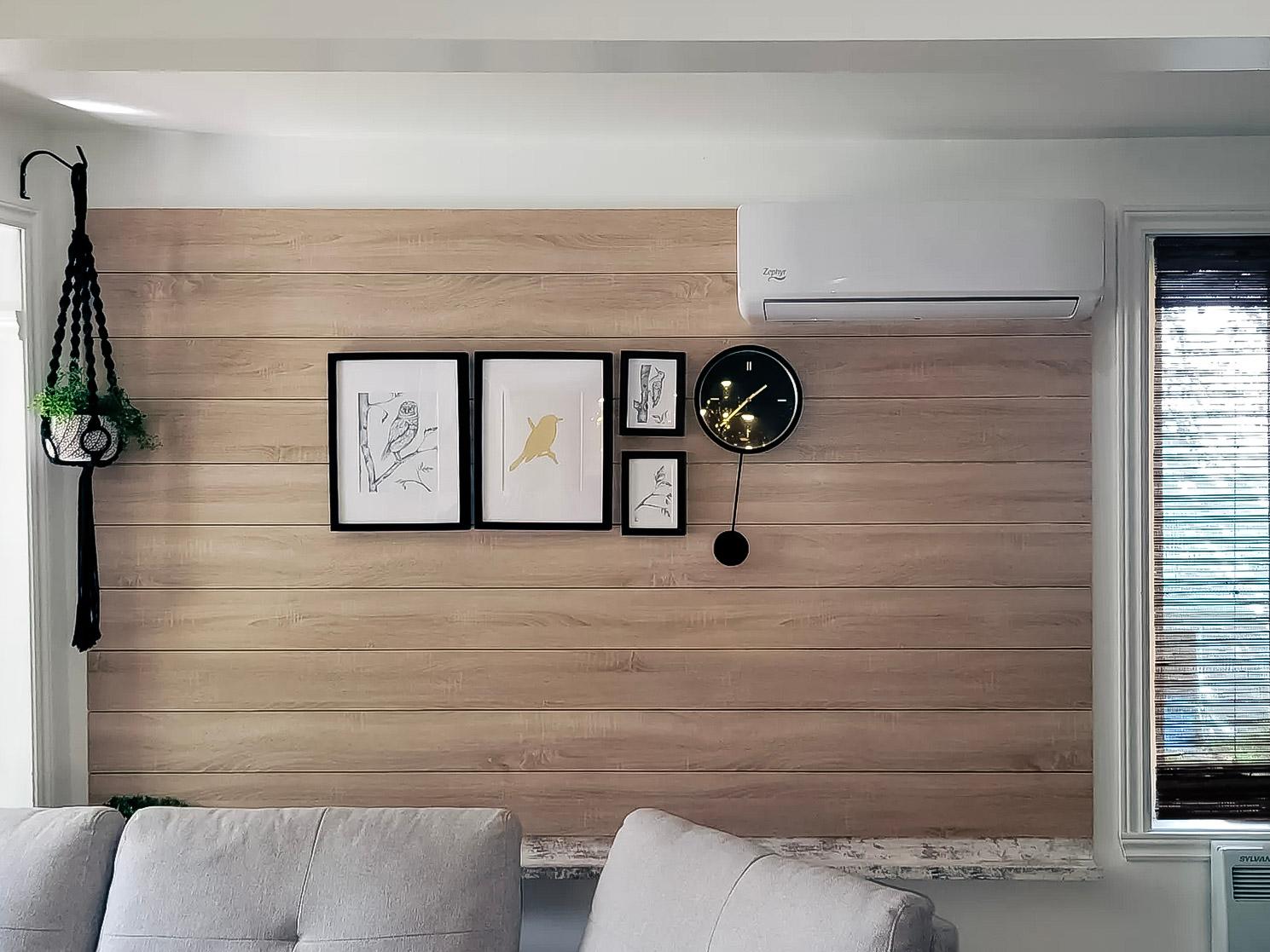 Mur en bois clair avec climatisation murale, galerie de cadres décoratifs, horloge design noire et plante suspendue dans un salon moderne.