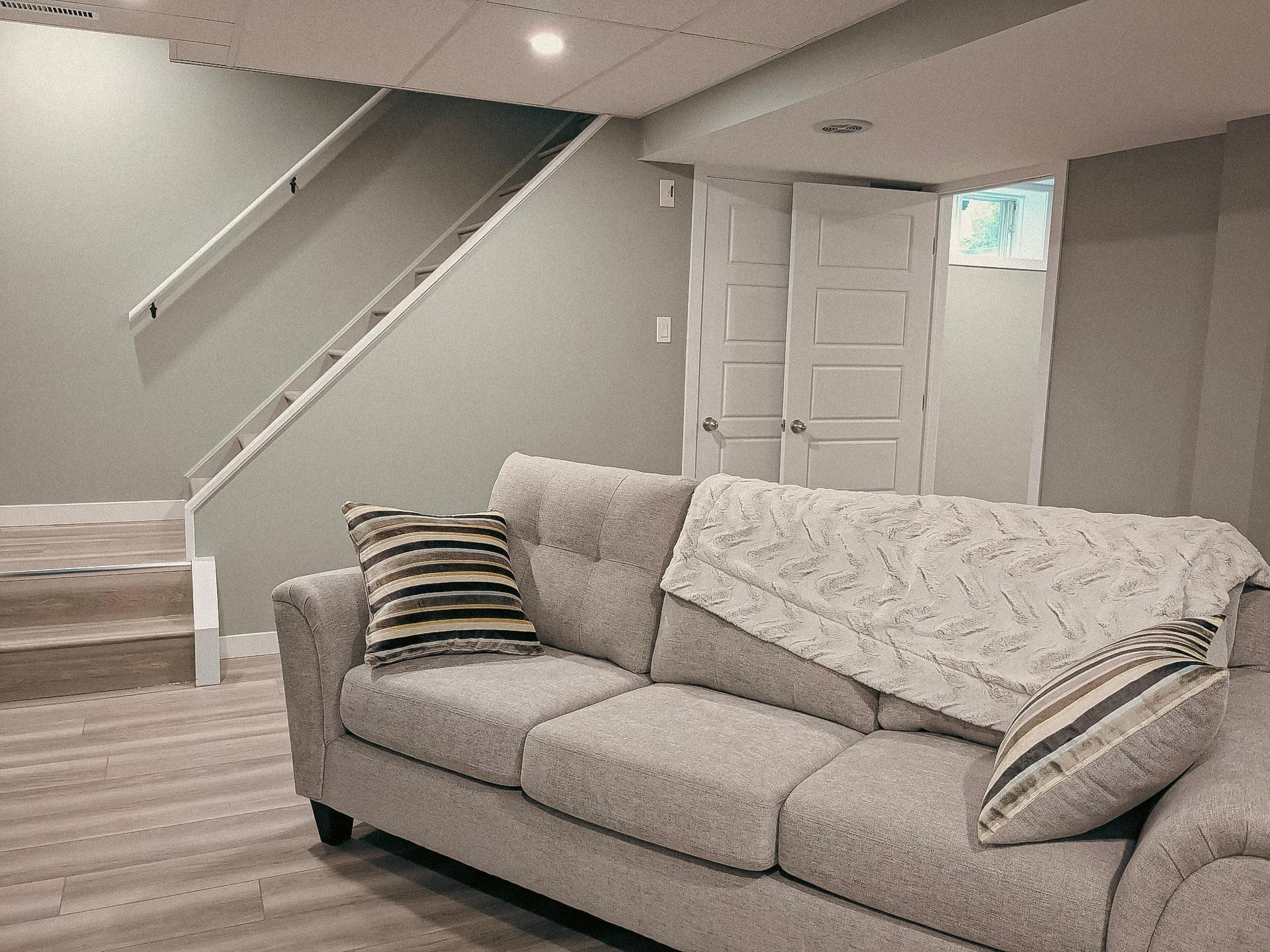 &nbsp;Sous-sol aménagé avec un canapé gris, des escaliers en bois et un mur neutre avec une porte blanche.