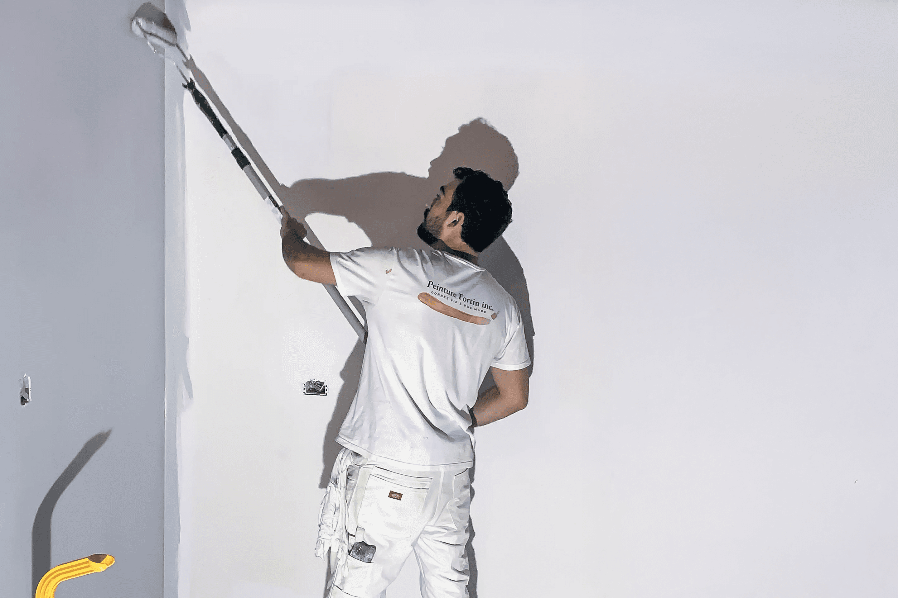 Peintre en bâtiment appliquant une couche de peinture blanche sur un mur à l’aide d’un rouleau télescopique, portant une tenue de travail blanche.