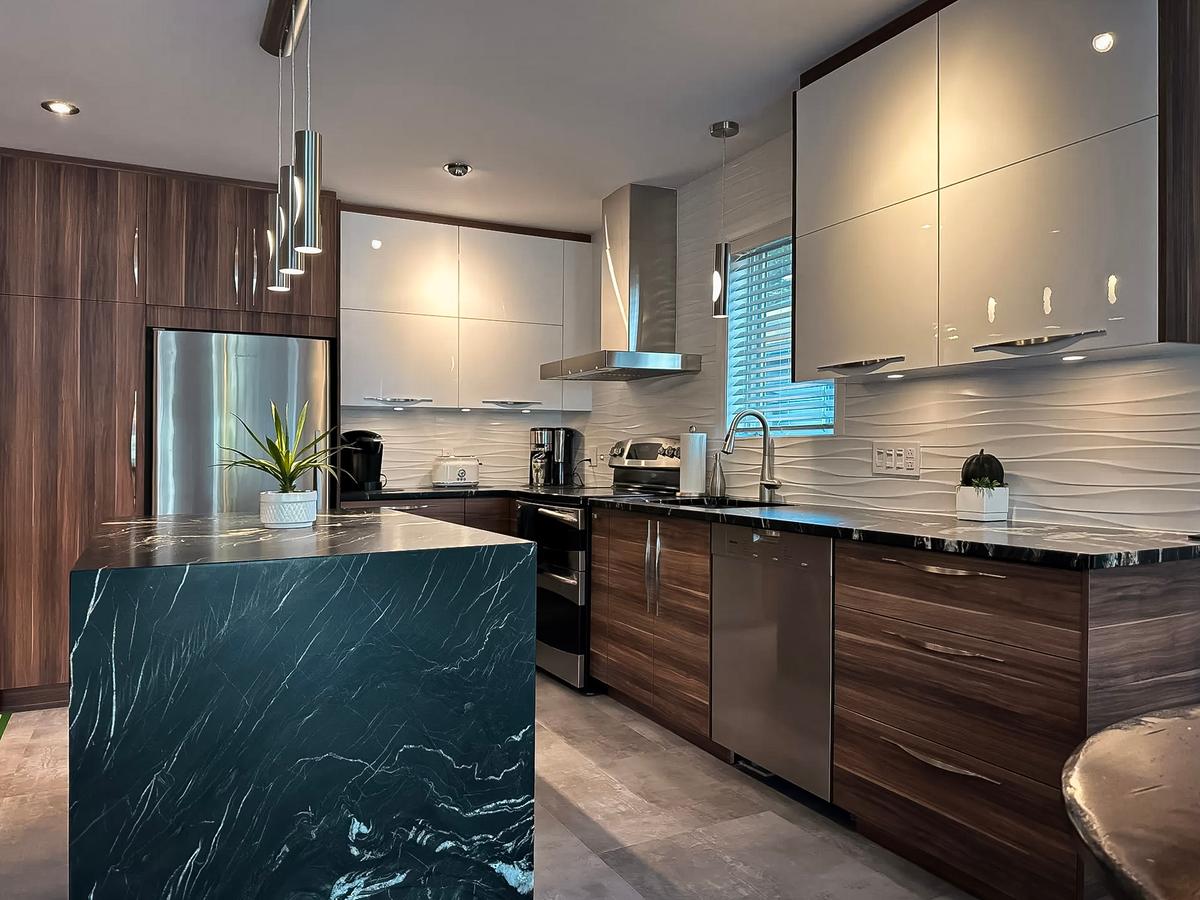 Cuisine moderne avec îlot en marbre noir, armoires en bois et blanc laqué, électroménagers en acier inoxydable et éclairage suspendu.