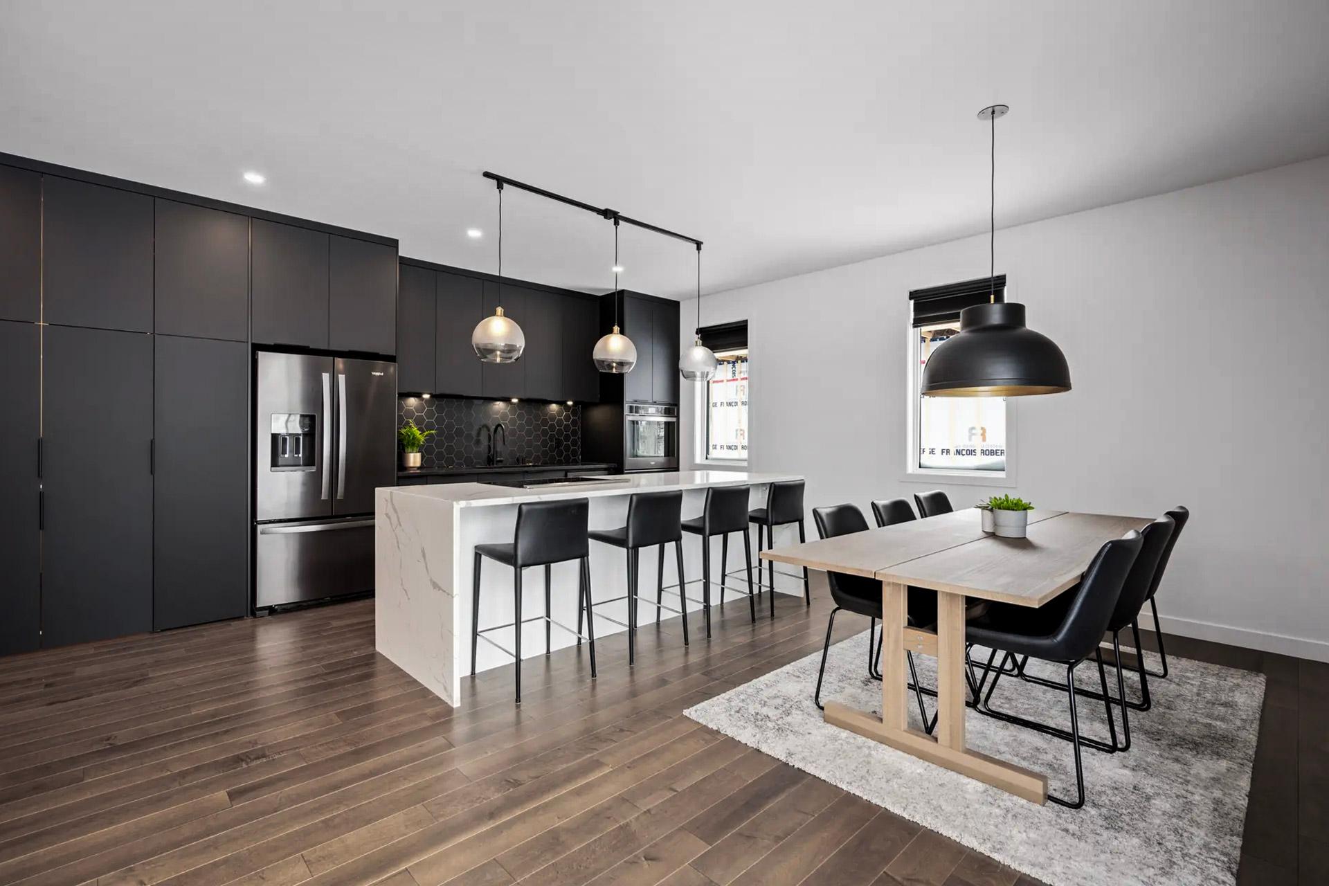 Cuisine moderne avec armoires noires, îlot en marbre et salle à manger élégante avec table en bois.