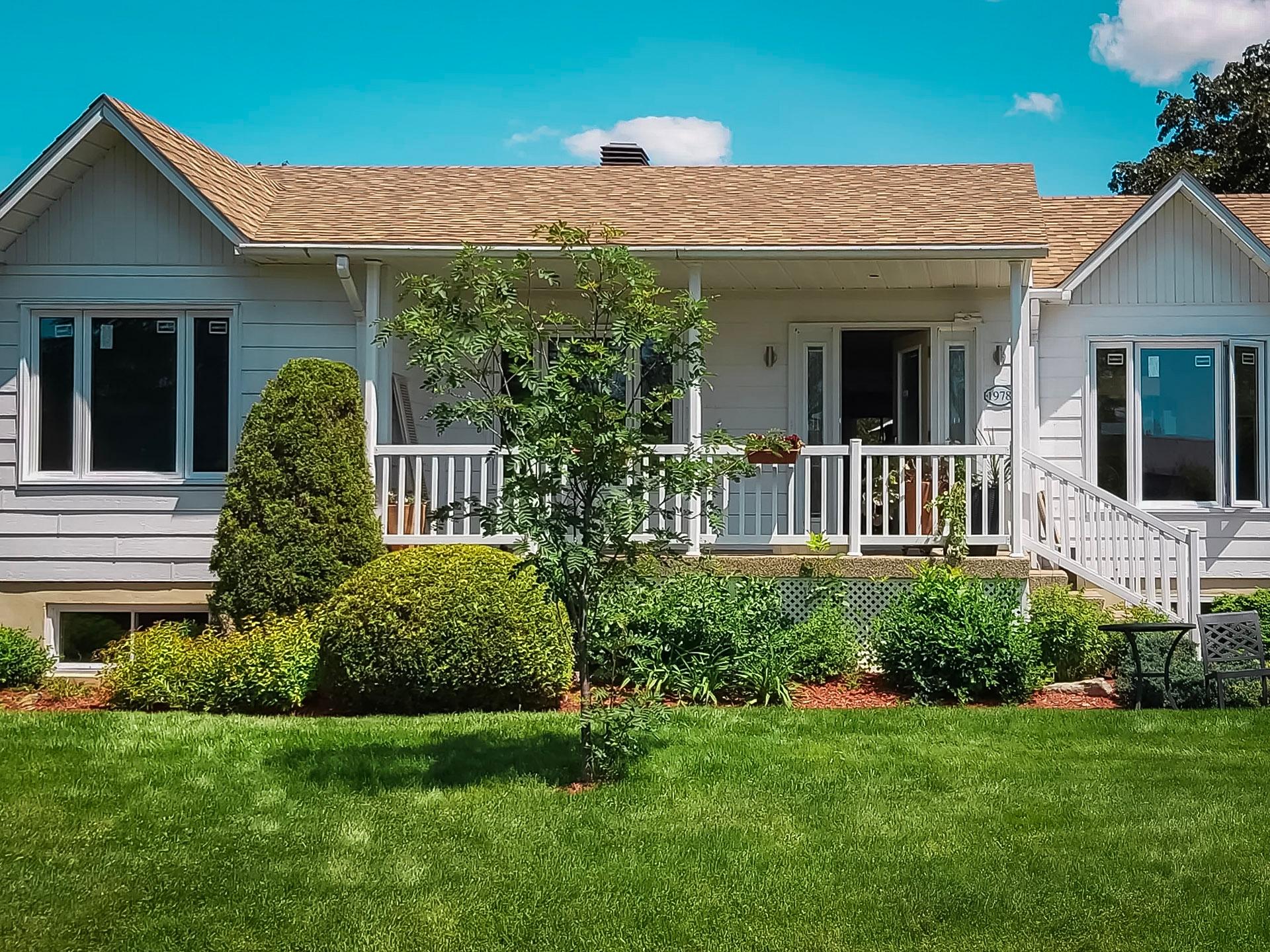 &nbsp;Maison de plain-pied avec terrasse en bois blanc, jardin paysager et pelouse verte sous un ciel bleu clair.