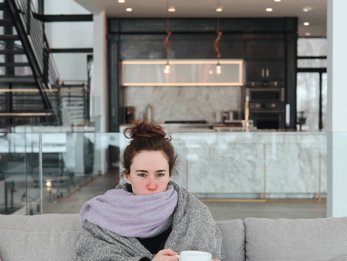 Femme emmitouflée dans une couverture et une écharpe, tenant une tasse chaude dans un salon moderne.