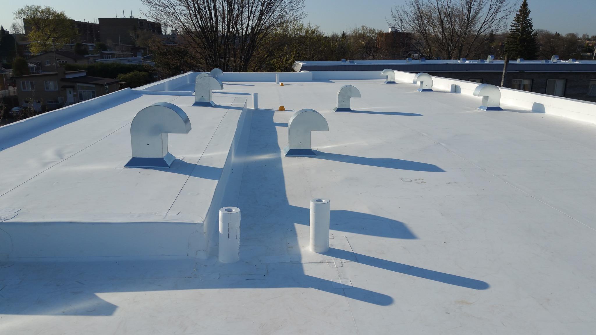 Toit plat recouvert d'une membrane blanche avec plusieurs évents métalliques et drains, assurant l'étanchéité et la ventilation d'un bâtiment résidentiel.