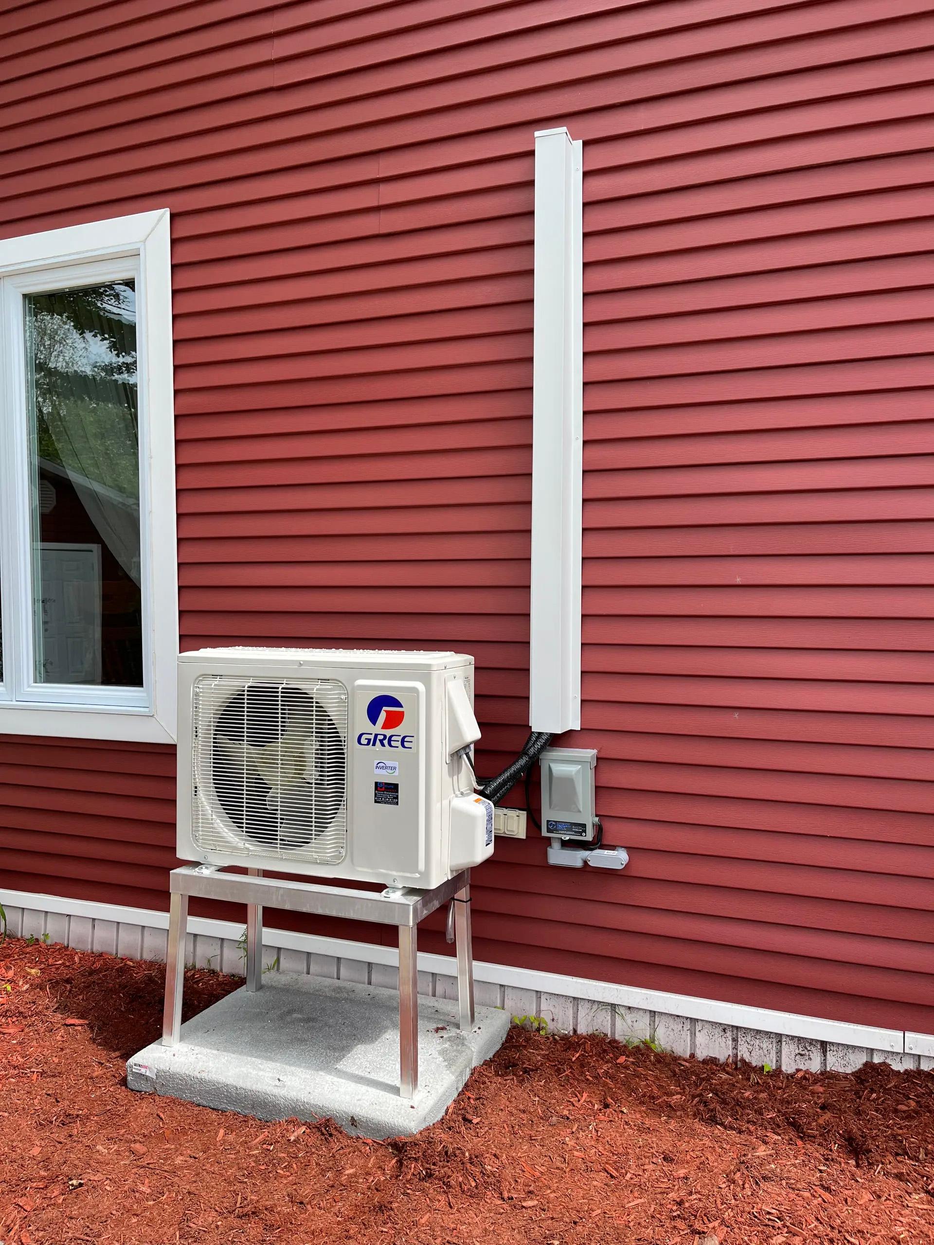 Unité extérieure de thermopompe Gree installée sur un mur de maison rouge avec boîtier de conduits et base en béton