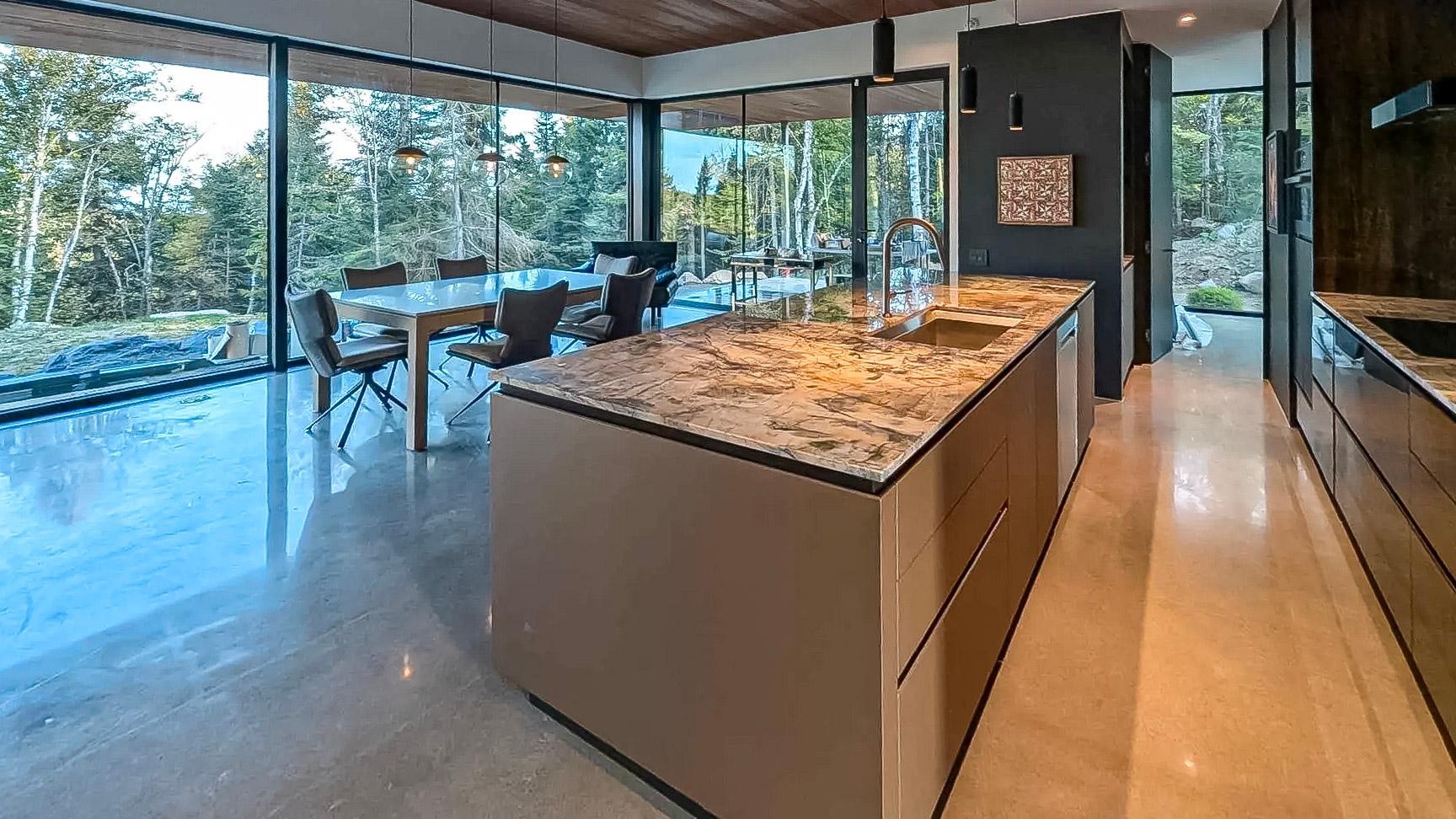 Cuisine moderne avec îlot central en marbre, grandes baies vitrées offrant une vue sur la forêt, plafond en bois et sol en béton poli.