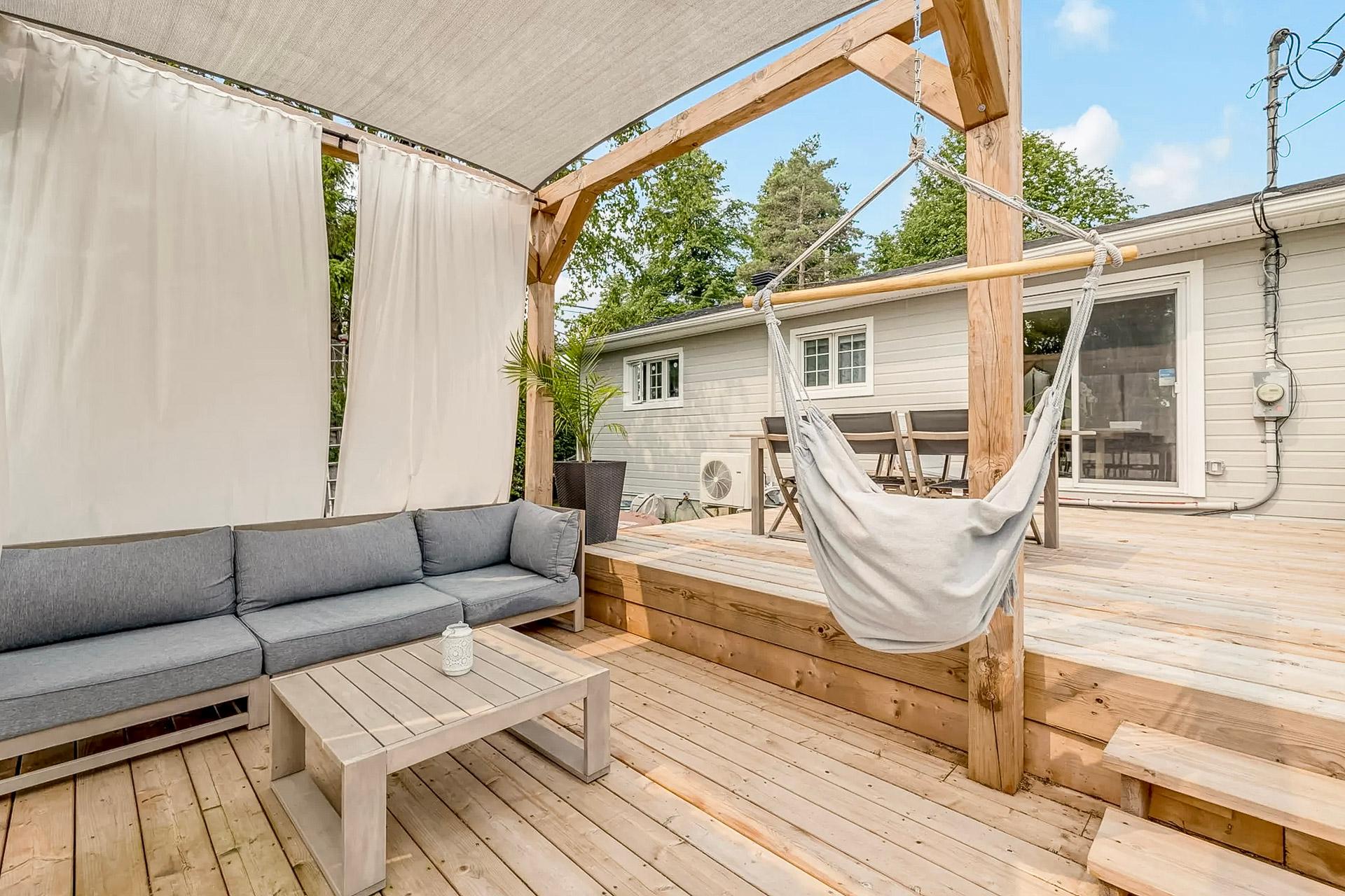 Terrasse en bois avec coin détente, canapé extérieur, rideaux blancs et chaise suspendue sous pergola