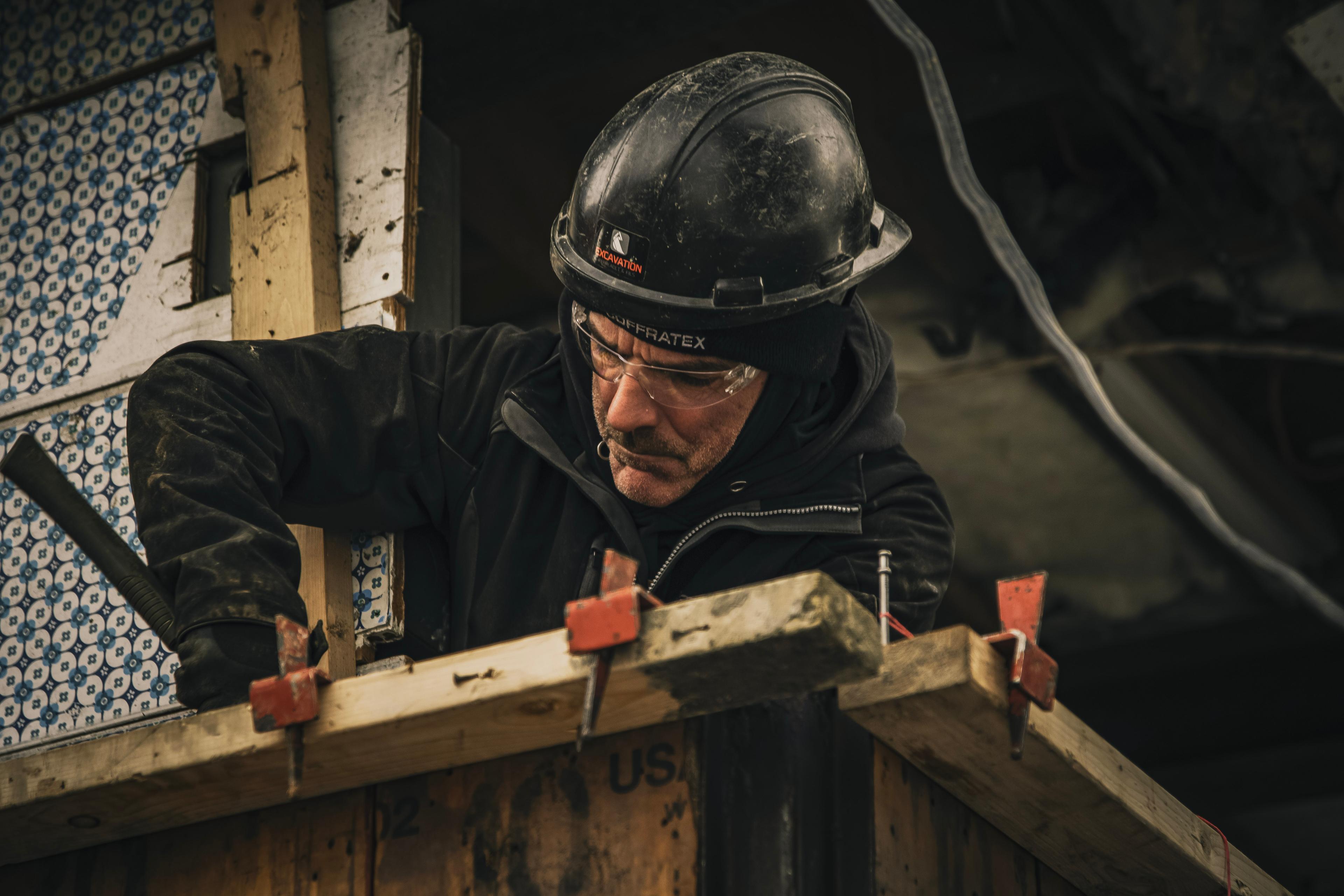 Travailleur portant un casque noir et des lunettes de protection, effectuant des travaux de menuiserie et de construction sur un chantier.
