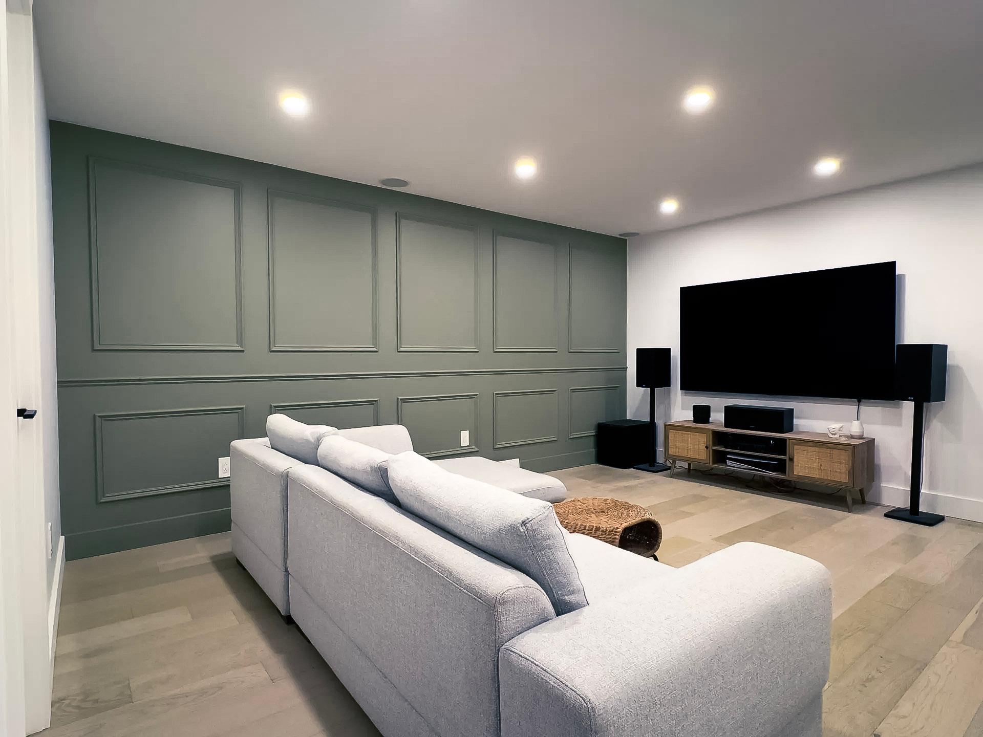 Salon moderne en sous-sol avec un mur accent vert, un grand canapé gris, un téléviseur écran plat et un système audio.